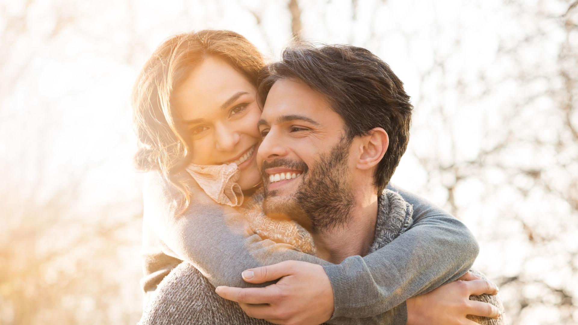 A smiling couple embracing outdoors in warm autumn sunlight, with bare trees softly blurred in the background